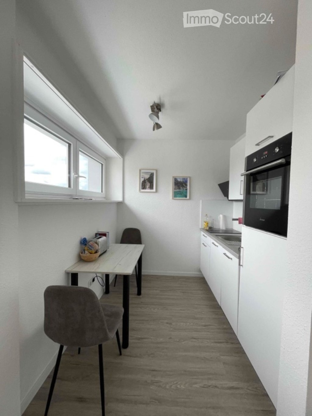 A modern, compact kitchen and dining area with white cabinets, a built-in oven, a wooden dining table, and two chairs. The space features light-colored walls, wooden flooring, and large windows allowing natural light to enter.