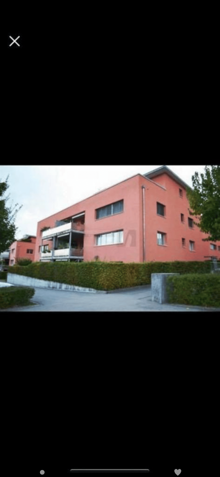 A modern, multi-story building with a pink facade and large windows, featuring a balcony and surrounding greenery.