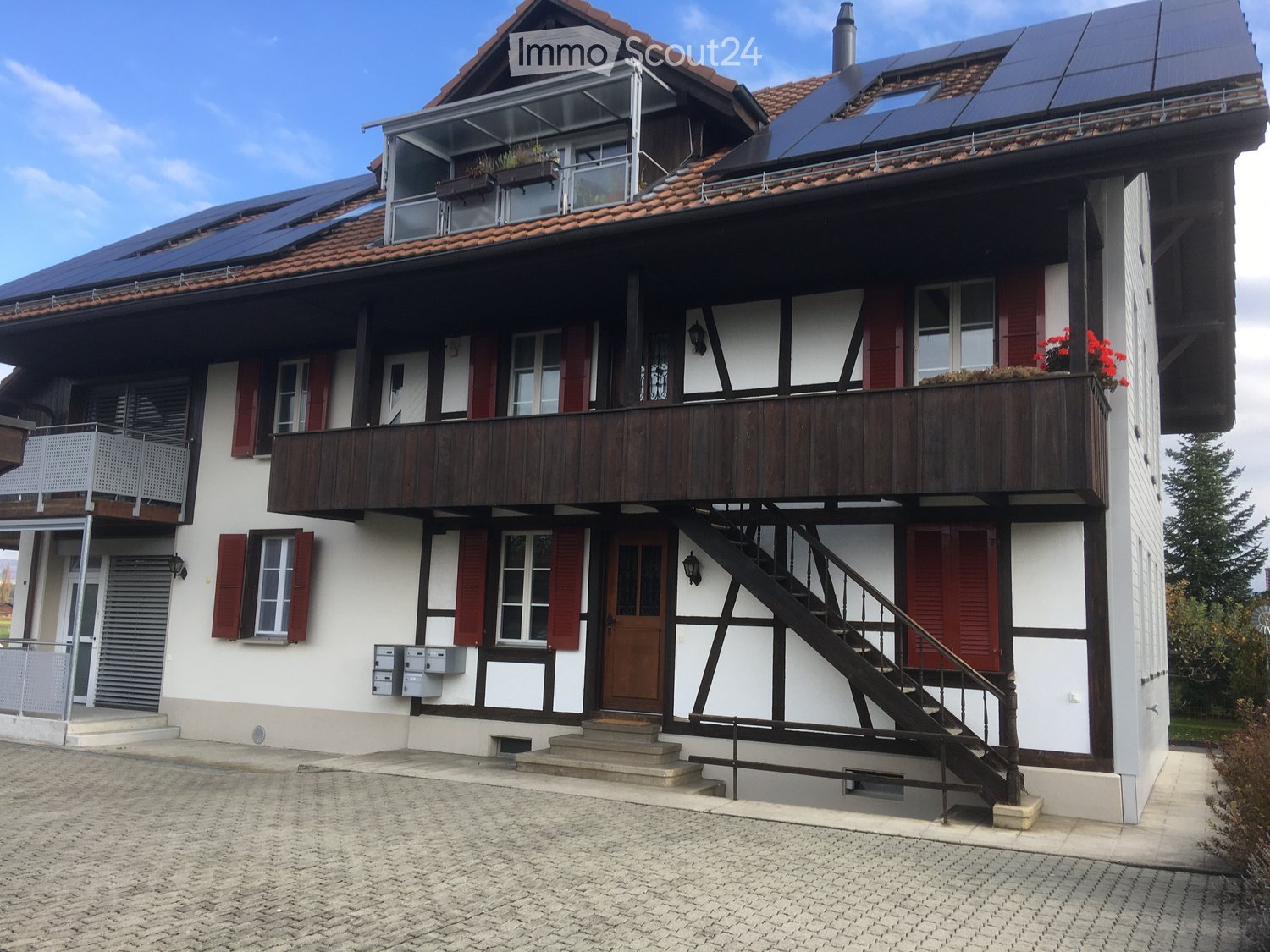 Two-story house with white walls, red shutters, wooden balcony, solar panels on the roof, and a staircase leading to the upper level.