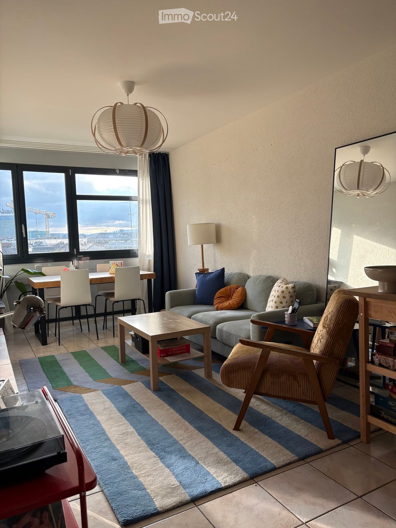 Modern living room with large windows, a grey sofa, coffee table, dining area, and decorative items.