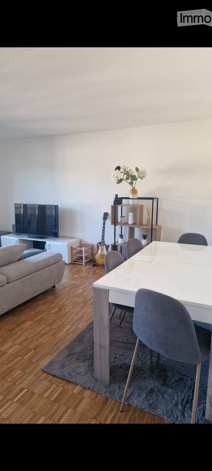 Modern living and dining area with a grey sofa, white dining table, grey chairs, a guitar, a shelf with decorative items, and a television.