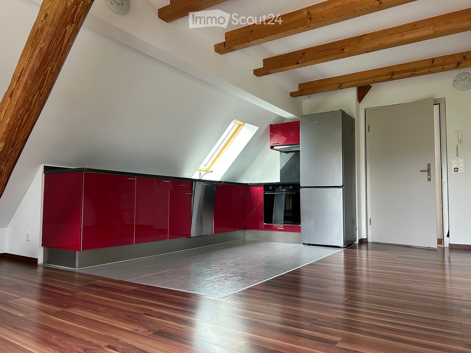 Empty kitchen with red cabinets, stainless steel appliances, wooden ceiling beams, and a window.