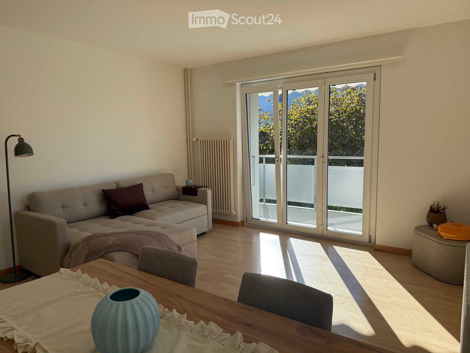 Bright living room with wooden flooring, large sliding glass doors leading to a balcony, a grey sectional sofa, a wooden dining table with chairs, and decorative items including a vase and a floor lamp.