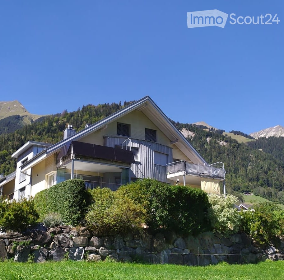 Two-story house with a balcony, surrounded by greenery and mountains in the background.