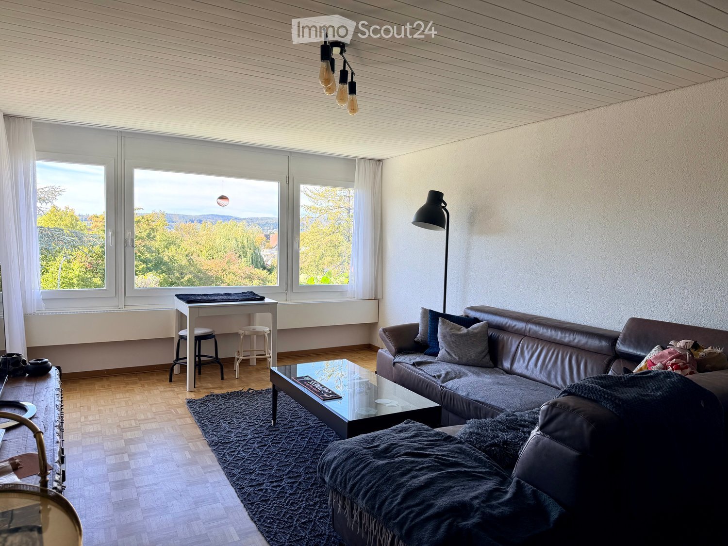 Modern living room with large windows offering a scenic view, featuring a dark leather sofa, glass coffee table, small table with stools, and a floor lamp.