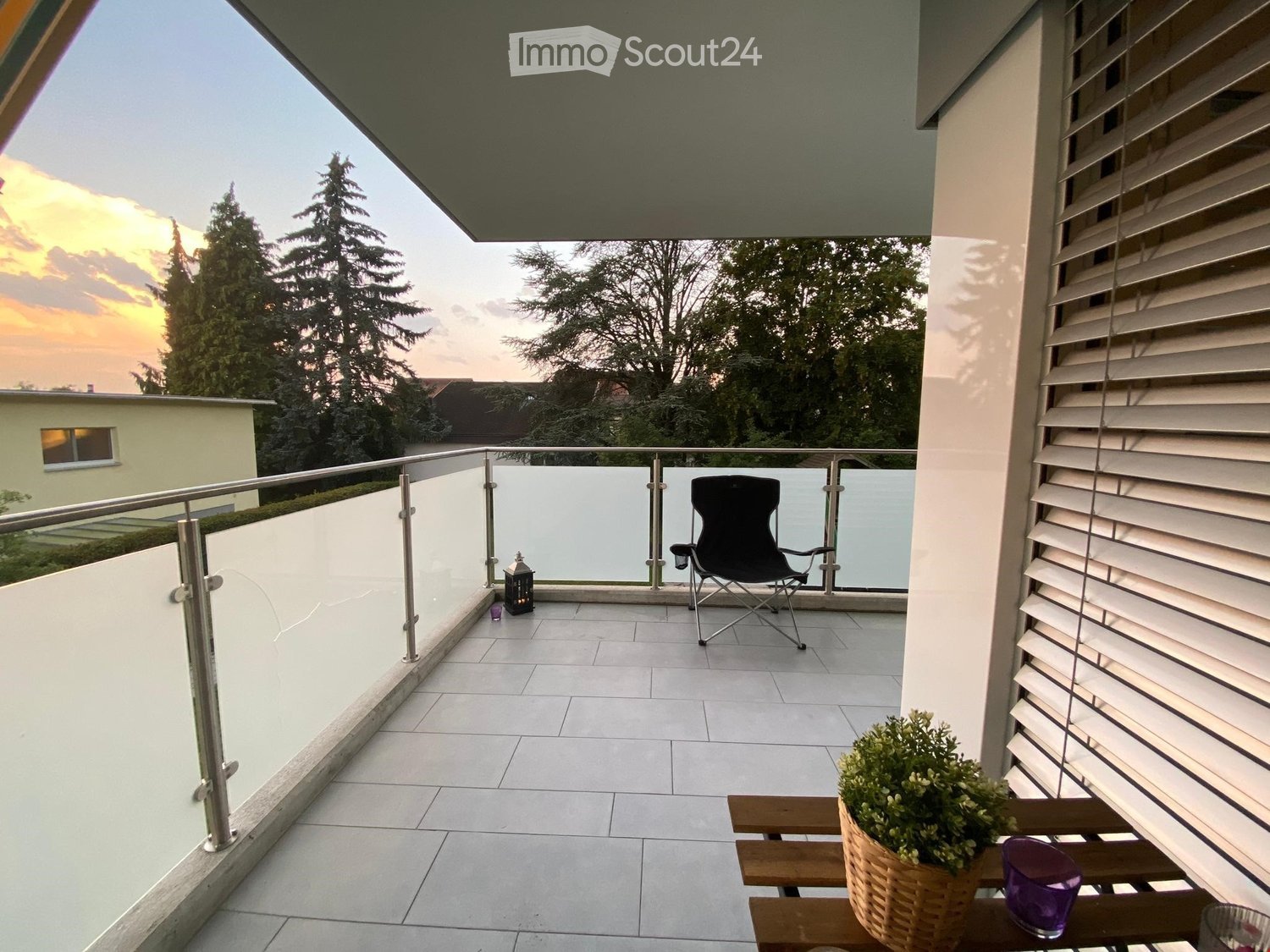 Balcony with tiled floor, glass railing, wooden table with a potted plant, black folding chair, and lantern.