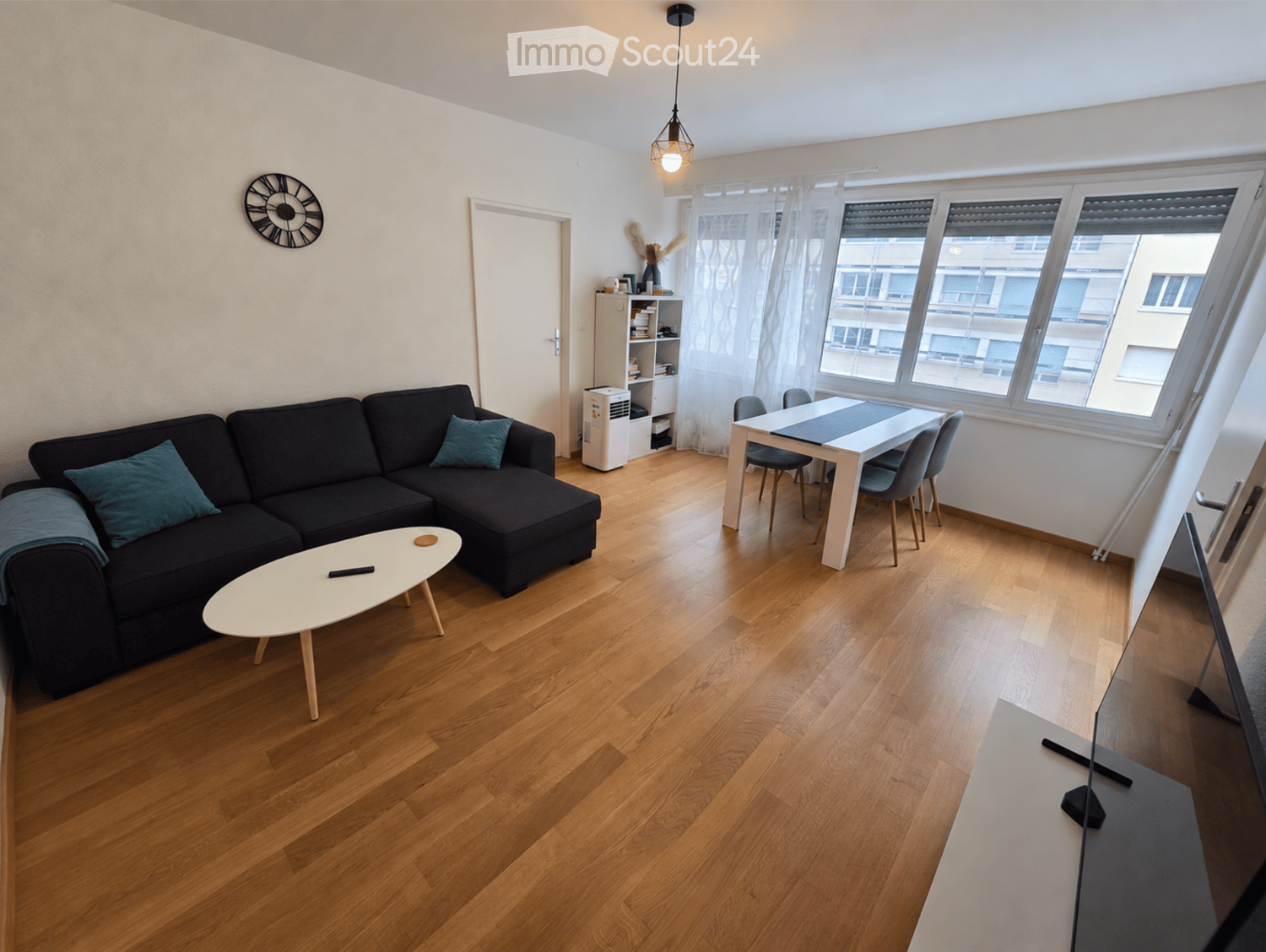 Modern living room with wooden flooring, a black sectional sofa, a white coffee table, a dining table with chairs, a screen, and a clock on the wall.