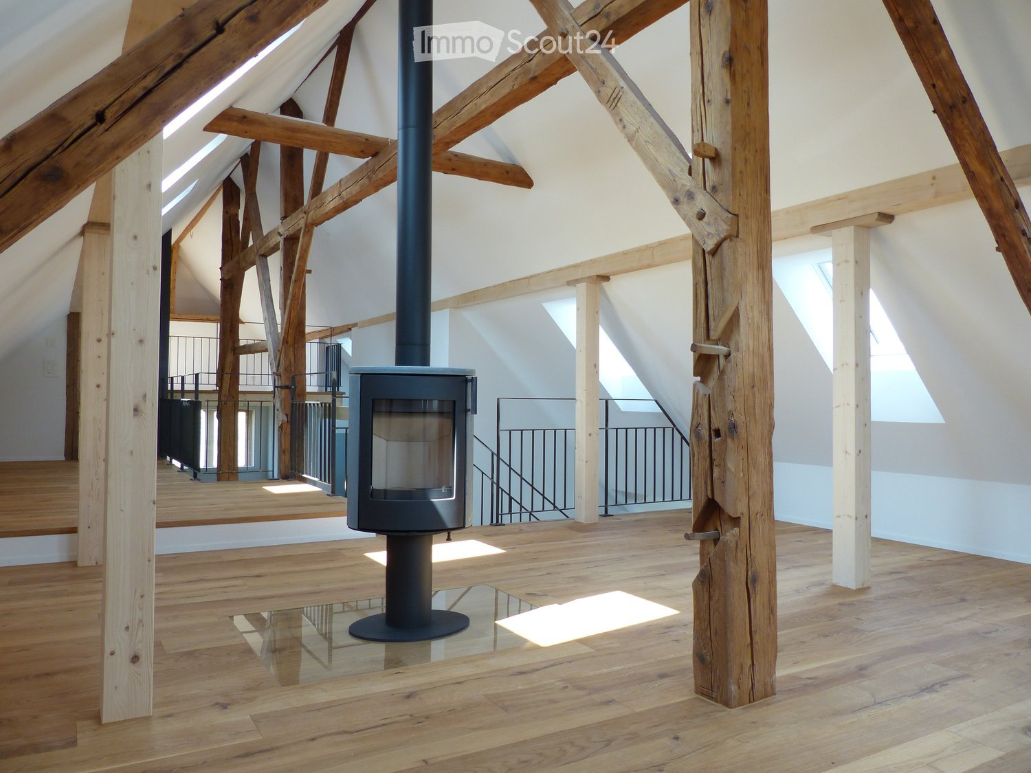 Open-concept attic space with exposed wooden beams, polished wooden floor, and a modern black freestanding fireplace.