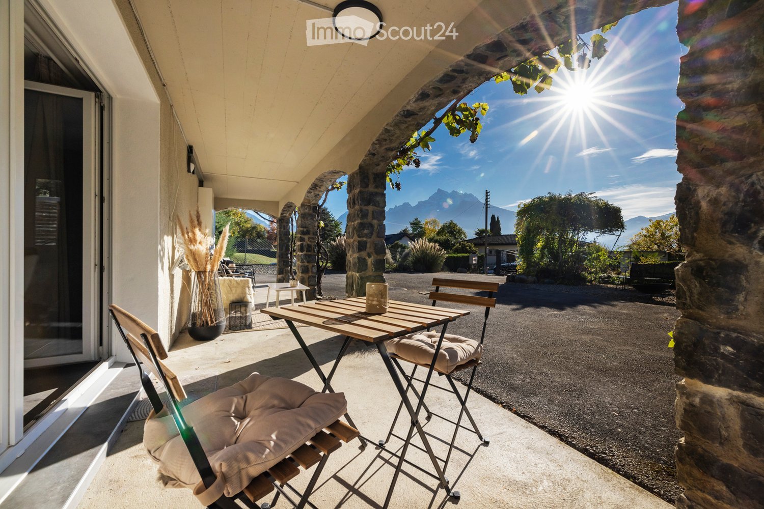 Balcony with a wooden table, two chairs, and a vase with dried plants, overlooking a scenic view of mountains and a road.