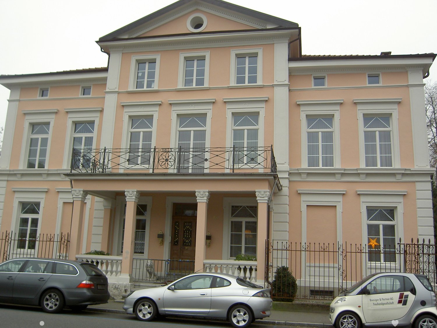 A two-story pink and white building with a balcony, ornate architectural details, and several cars parked in front.