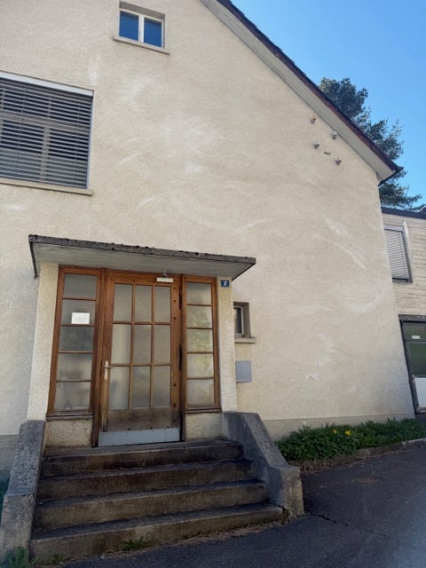 A beige building with a wooden door and glass windows. The building has a slanted roof and a small balcony on the second floor. The building has a number 7 on the wall above the door.