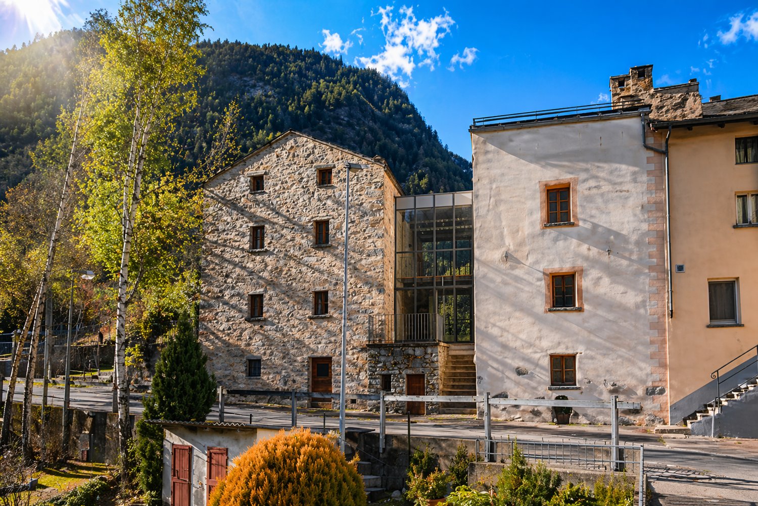 Two-story stone building with glass extension, multiple windows, surrounded by trees and mountains