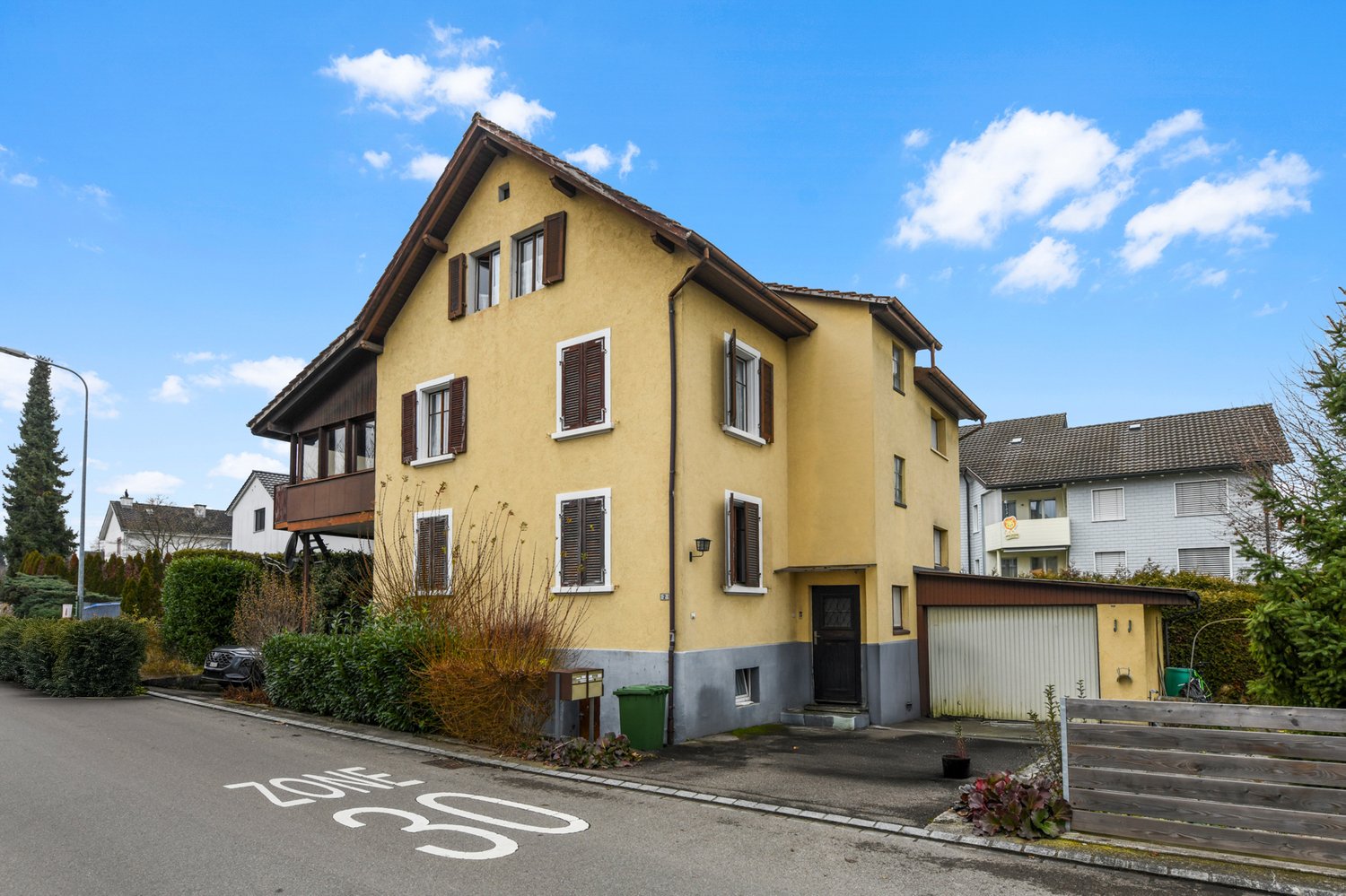 A three-story house with yellow walls and brown trim, several windows, a black front door, a garage, and an entrance with stairs. There are also a wooden fence and a green trash bin on the property.