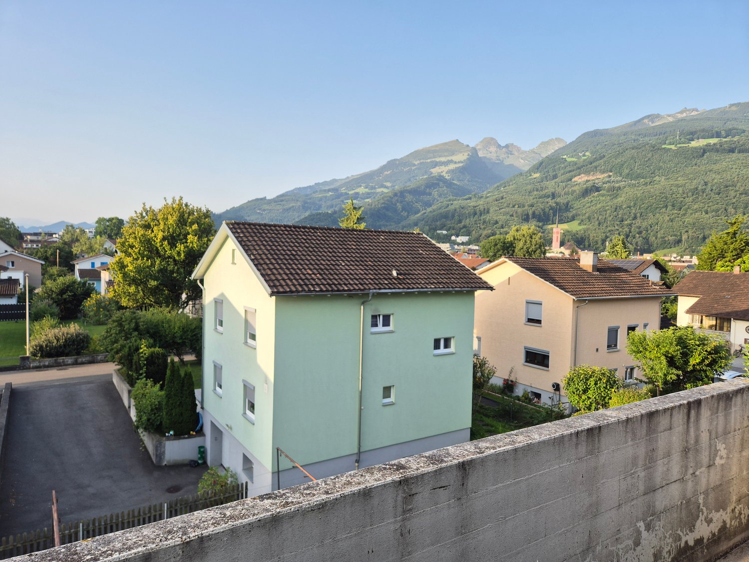 Two-story house with brown roof, green walls, several windows, located in a neighborhood with other houses, trees, and mountains in the background. The road and parking area are in front of the house.