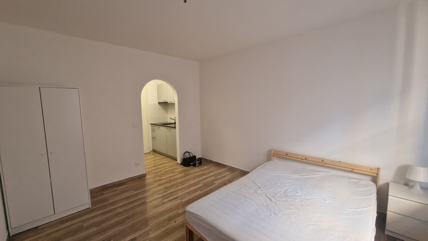 The image shows a studio apartment with a bed, a wardrobe, and an arched doorway leading to a kitchenette area. The room has hardwood flooring and white walls. A washing machine is visible in the kitchenette area.