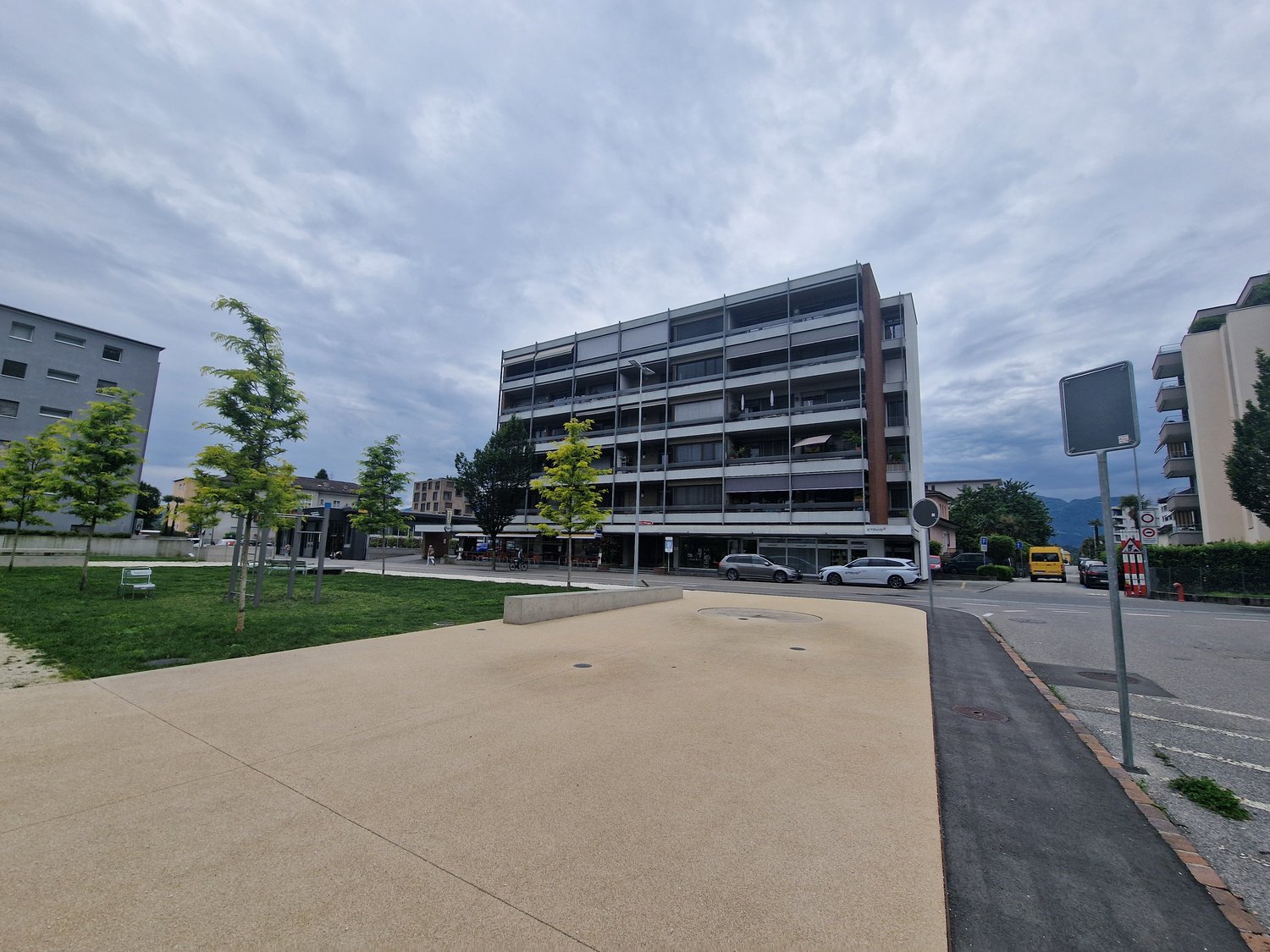 A multi-story building with many windows, surrounded by a grassy area and trees, with a paved walkway and streetlights.
