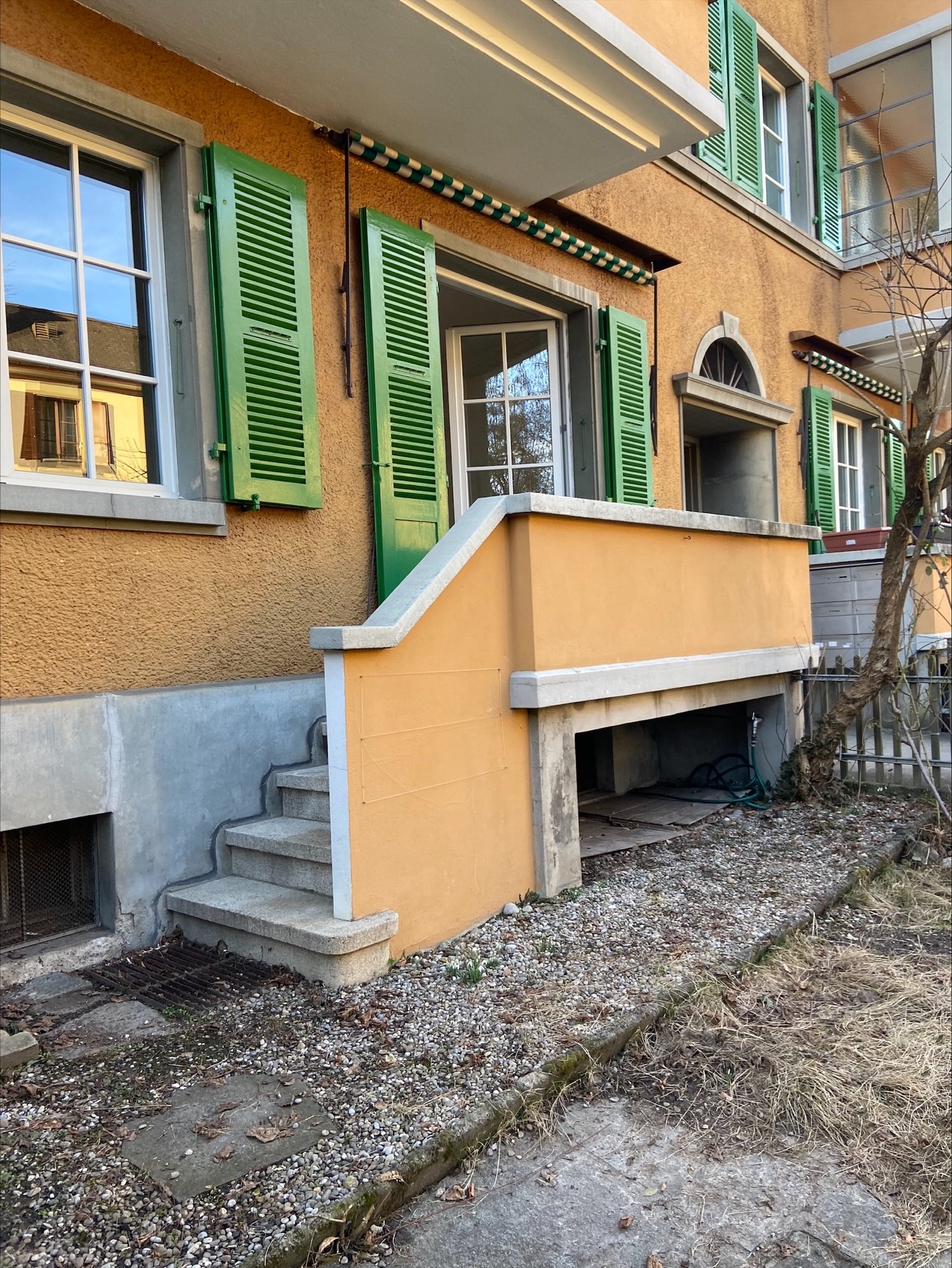 2-story building with yellow exterior walls, green shutters, and a balcony on the upper level. There are stairs leading up to the entrance and a small patio area in the front.