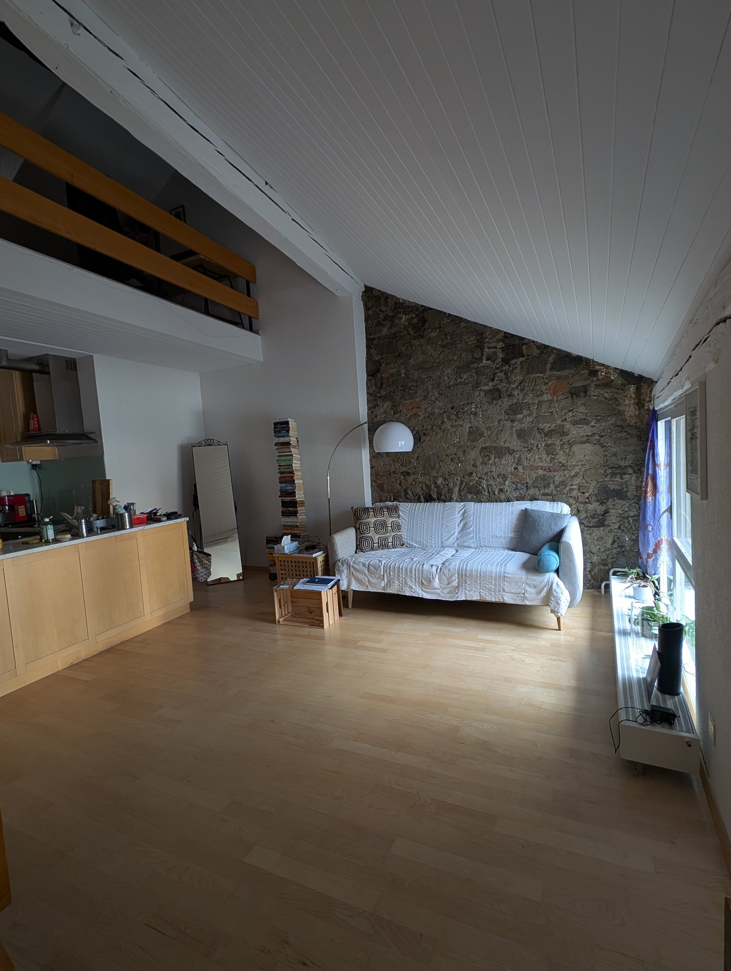 modern room with wooden floors, white walls, large sofa, desk with books, mirror, balcony, window with curtain