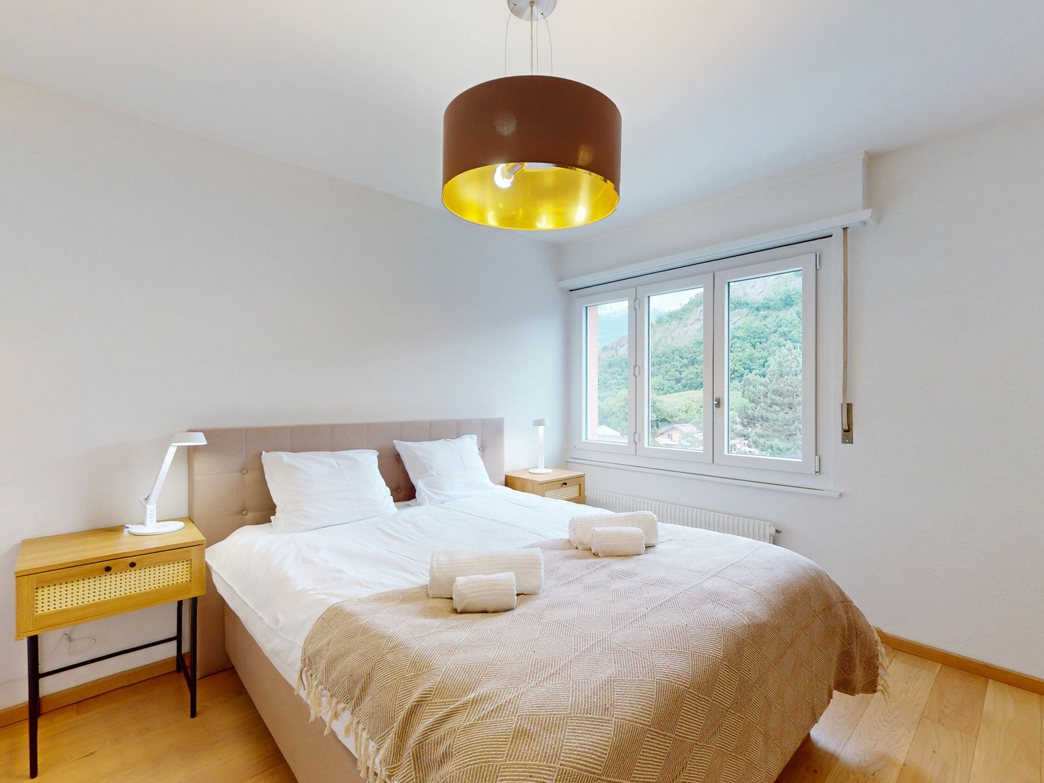 Spacious bedroom with large windows offering a scenic view, a double bed with white bedding and a beige patterned blanket, a wooden nightstand, and a pendant light fixture with a brown and yellow shade.