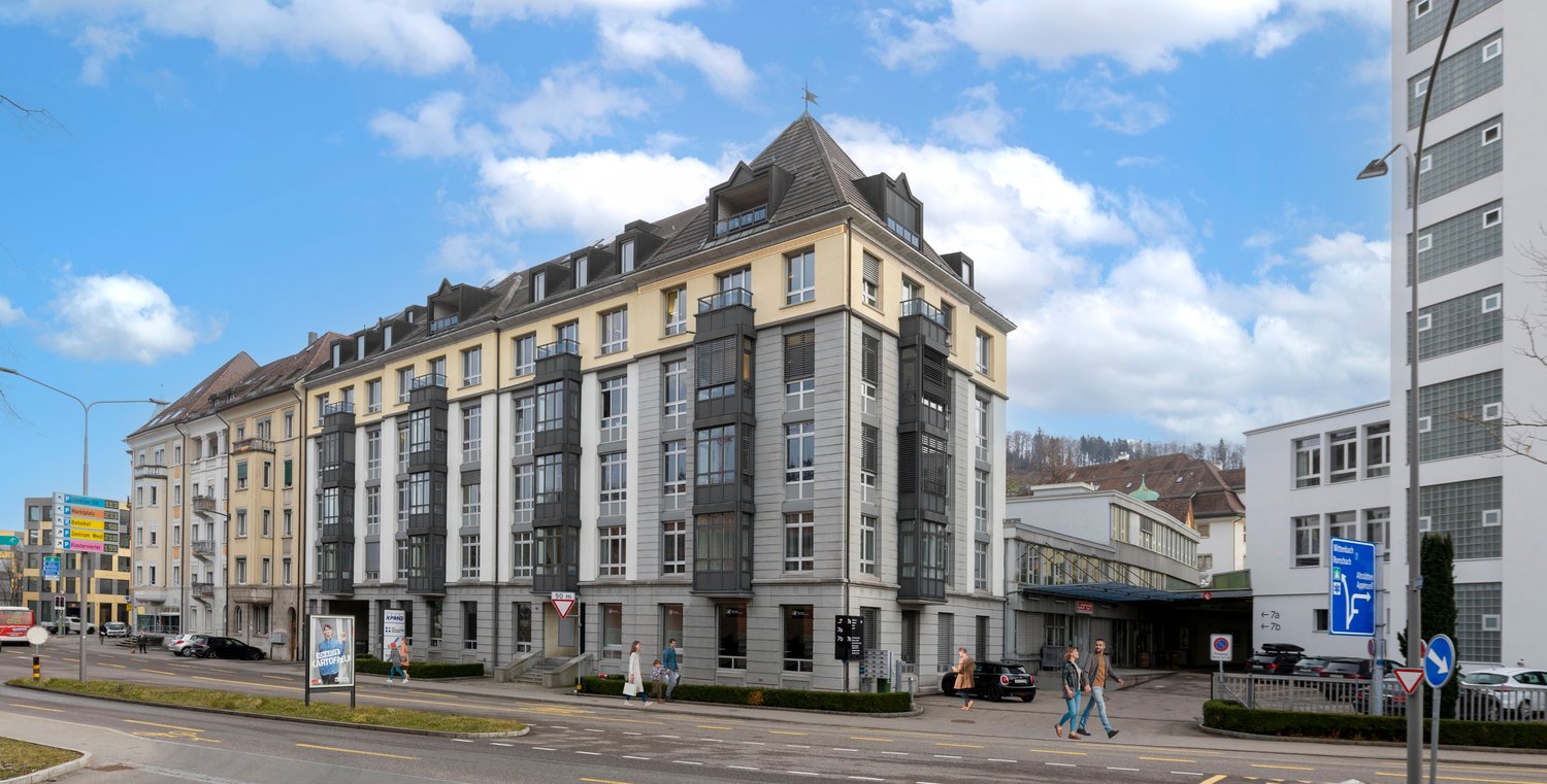 Multi-story building with a mix of residential and commercial spaces, featuring a distinctive roofline with dormers and a tower, balconies, and large windows. The building has a yellow and gray color scheme and is situated on a street with other buildings