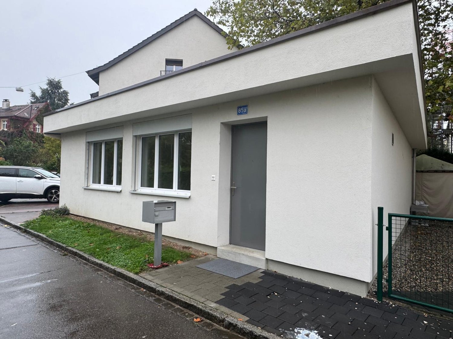 Raised ground floor, white exterior, 3 windows, gray door, gray mailbox, paved walkway, green fence, grass lawn, single car parked in the street