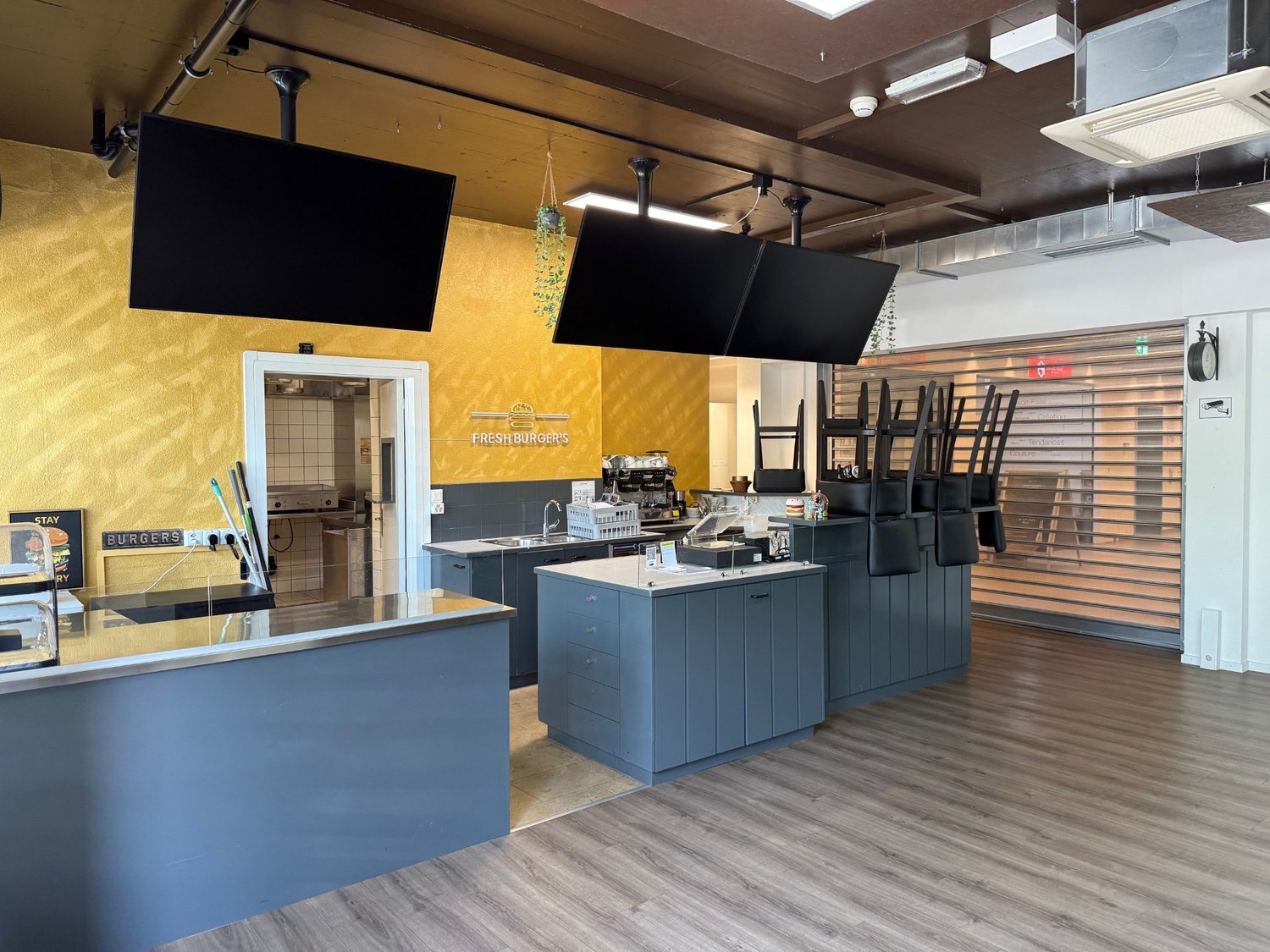 The image shows the interior of a restaurant or cafe. The space has a modern and industrial design, with yellow walls, black countertops, and a gray tiled floor. There is a counter area with a Fresh Burgers sign, and the kitchen area is visible with vario