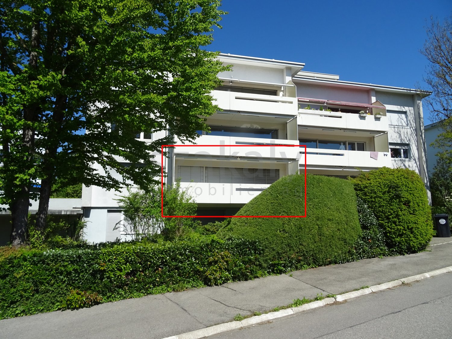 2-story apartment building, white exterior, second floor balcony, red framed windows, hedges, trash bin on sidewalk