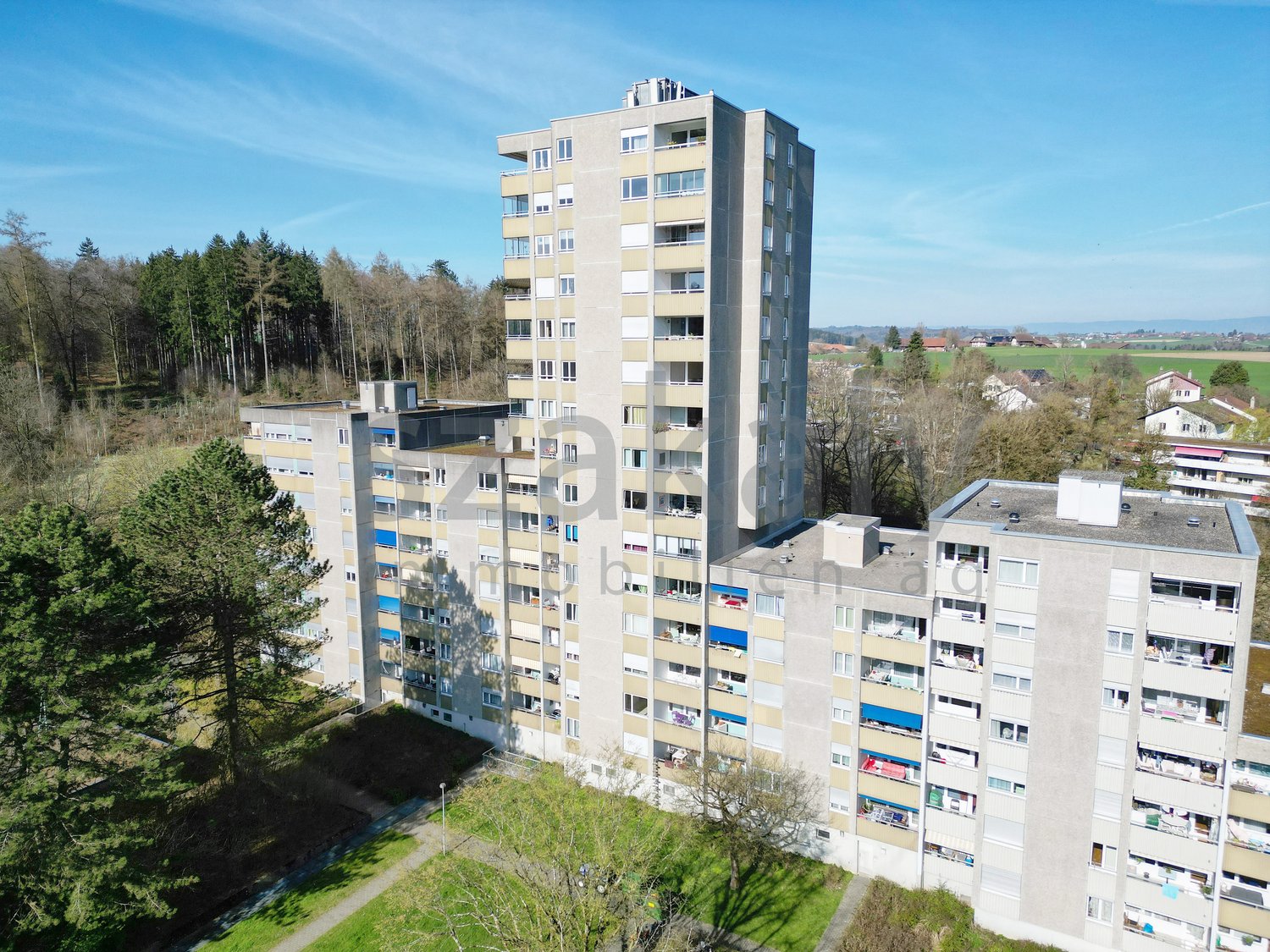 Aerial view of multi-story apartment building, surrounded by trees, several houses in the background, some with green roofs.