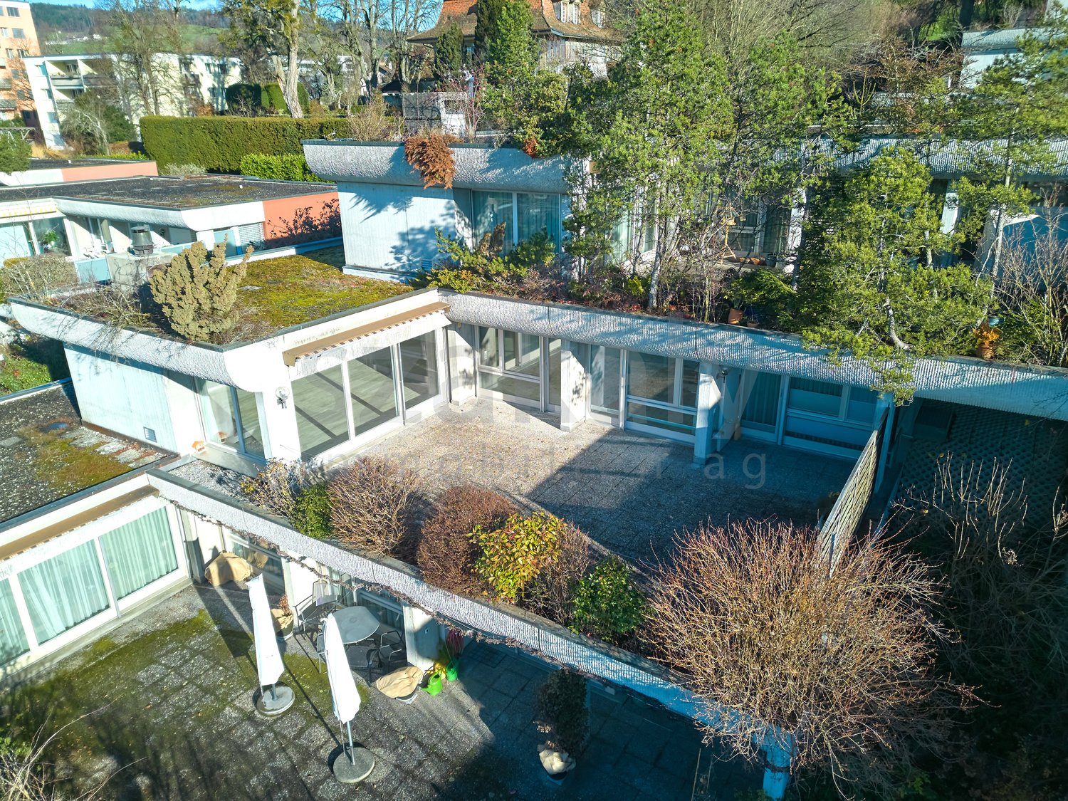 Modern house with a rooftop terrace, surrounded by trees and plants, open patio with outdoor furniture, multiple windows.