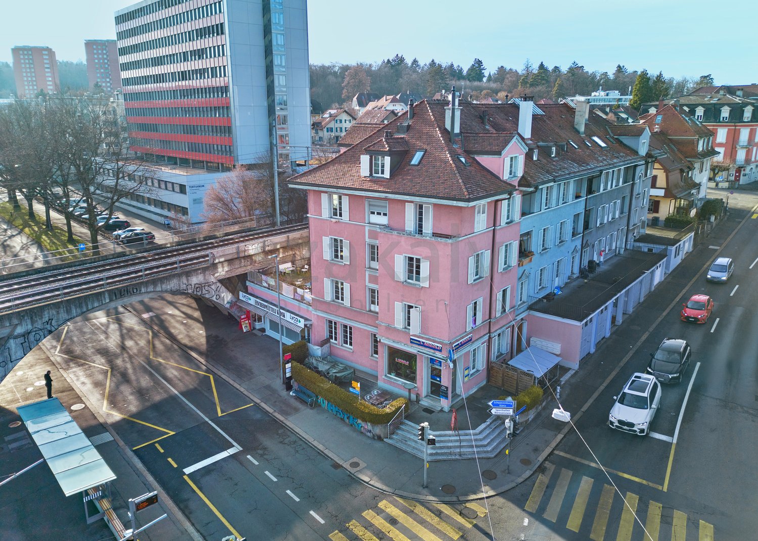 Aerial view of a three-story building, pink facade with many windows, ground-level entrance, street with cars parked on both sides.