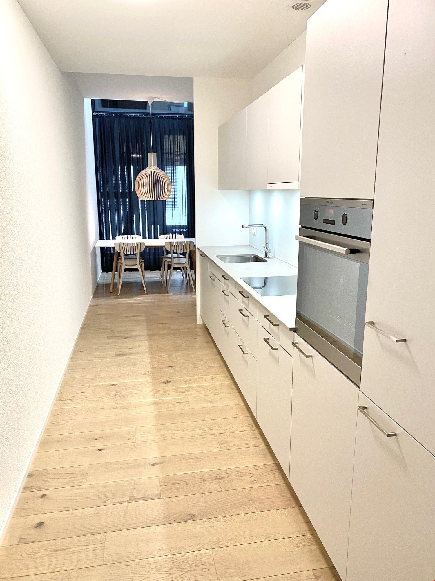Modern kitchen with white cabinets, stainless steel appliances, and a wooden dining table with chairs visible through the doorway