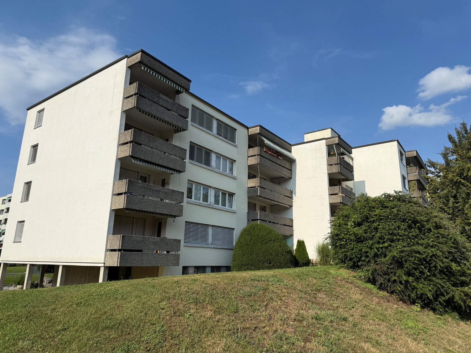 multi-story building, white exterior, balconies, set on a hill, surrounded by greenery
