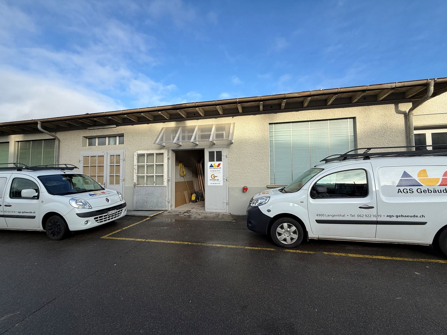 Single story building with a brown roof, white walls, several windows, and two vans parked in front