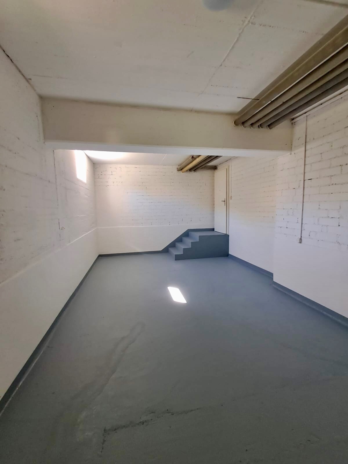 Empty room, white brick walls, concrete floor, stairs leading to a raised area, pipes on the ceiling, windows