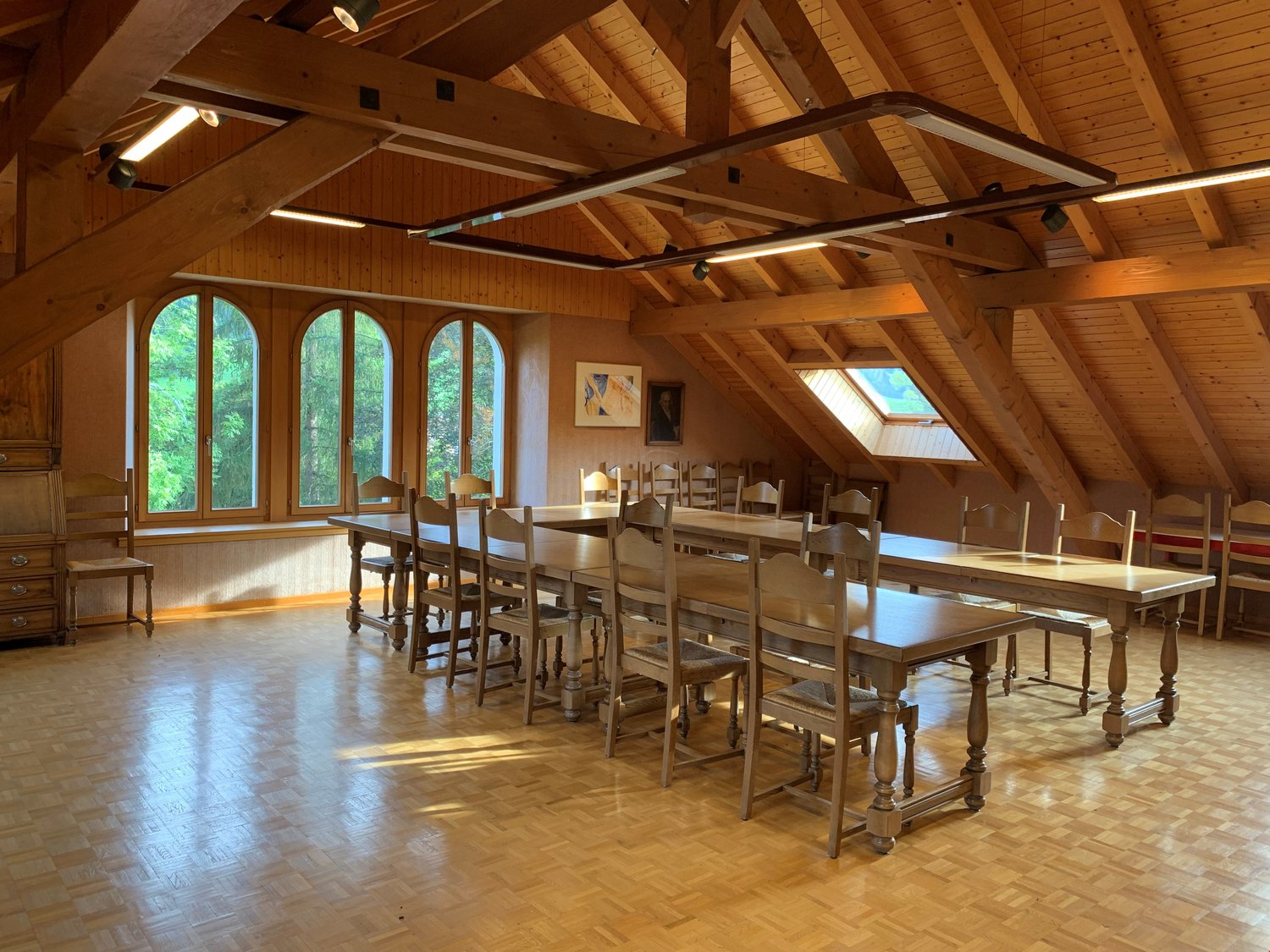 Large open-concept dining room with wooden beamed ceiling, arched windows overlooking the surrounding forest, wooden tables and chairs, and hardwood flooring.