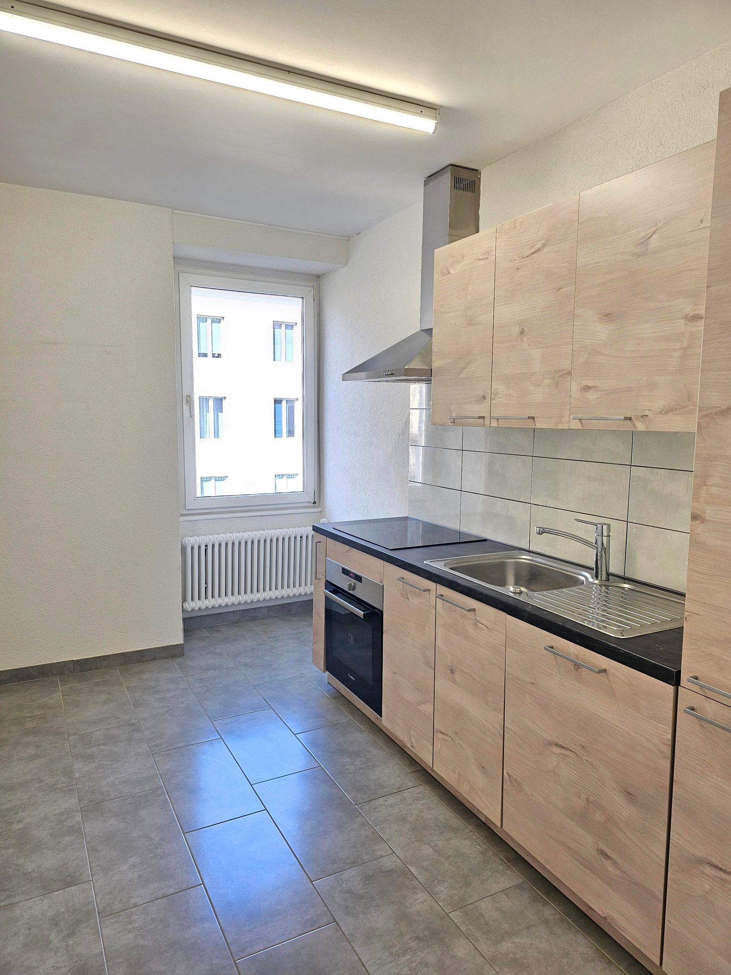 kitchen with light gray tiles, black countertop, wooden cabinets, silver sink, extractor hood above the stove, window with white radiator, and a white ceiling light