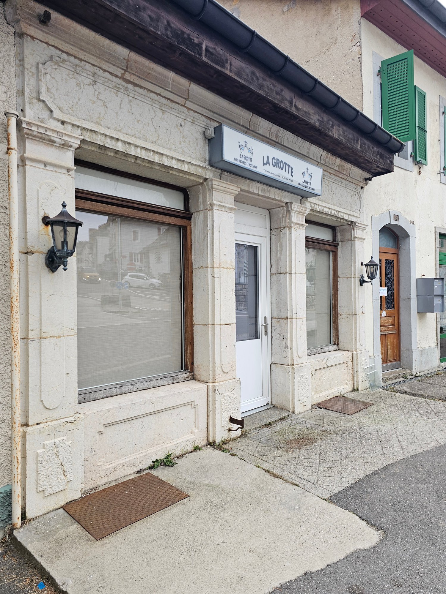 stone-made building with white facade, 3 large windows, entrance with glass door, lamp mounted on the wall
