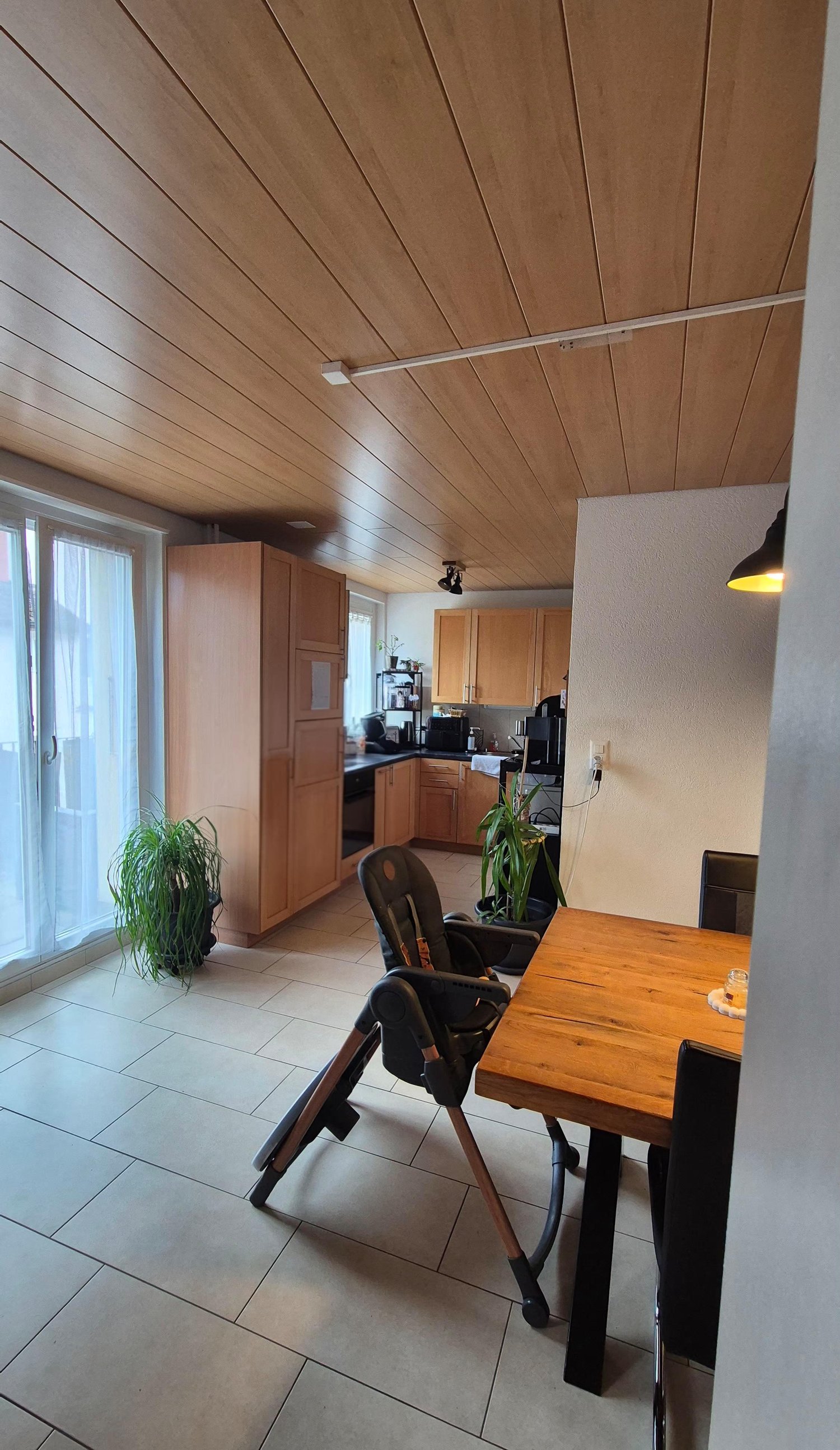 Dining area with wooden table, chairs, and a high chair. Wooden cabinets, kitchen appliances, and a window with curtains. Potted plants on the floor.