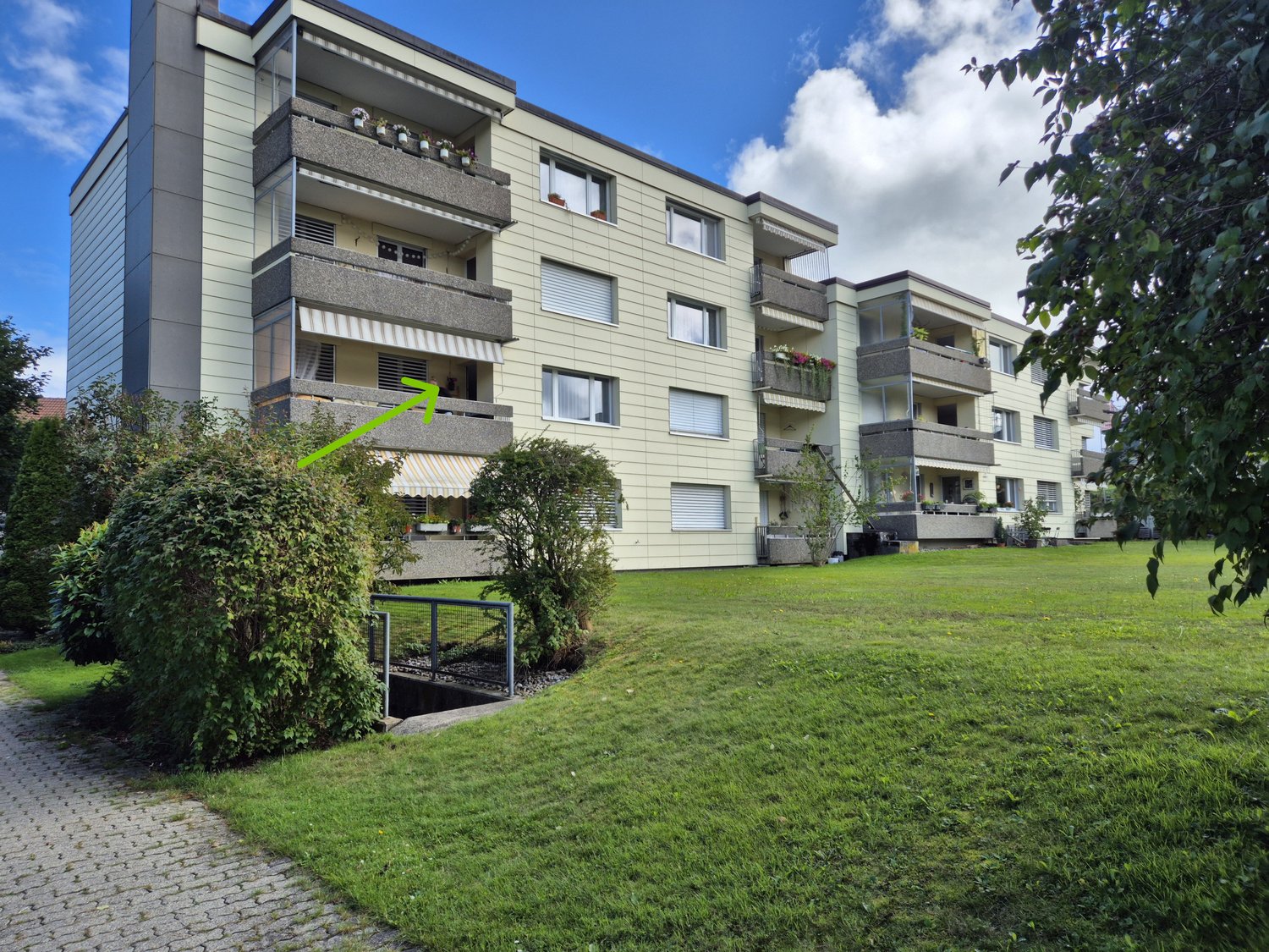 Apartment building, beige facade, multiple balconies, green garden in front, trees, grassy area