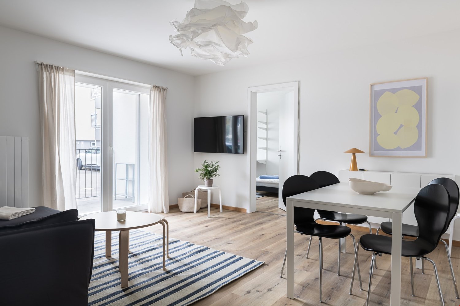 Living room with white walls, wooden floor, glass door to balcony, TV mounted on wall, dining table with 4 chairs, small side table with plant, white rug, lamp, picture frame on wall