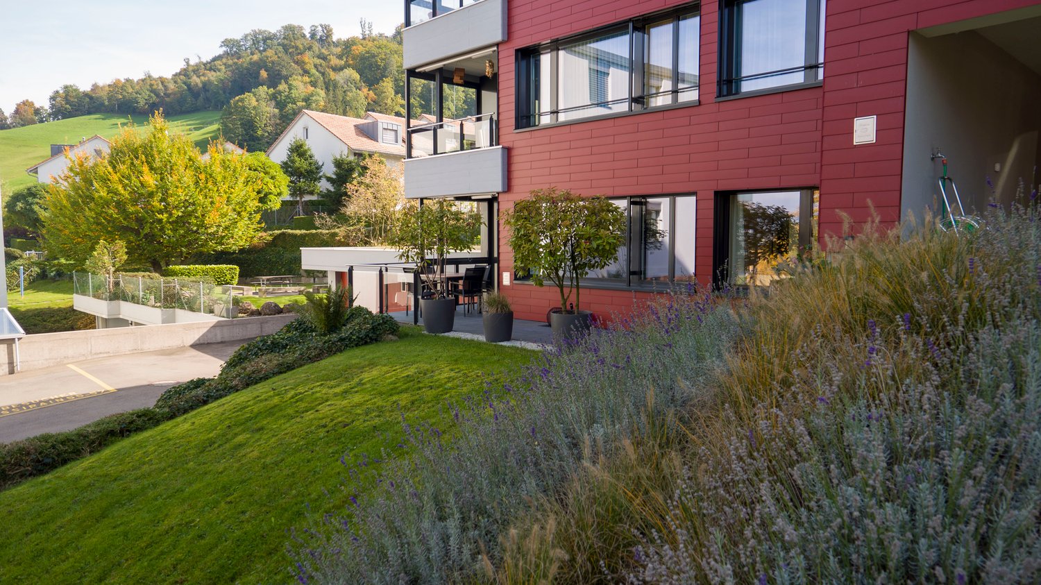 Two story house, red brick, white windows, balcony with fence, paved terrace, garden with green lawn, trees, shrubbery