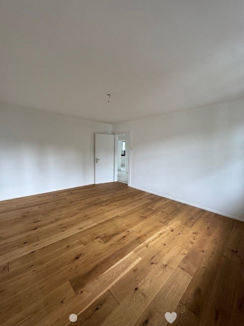 Empty room with white walls and hardwood flooring. The room has a door visible and appears to be a living or bedroom space.