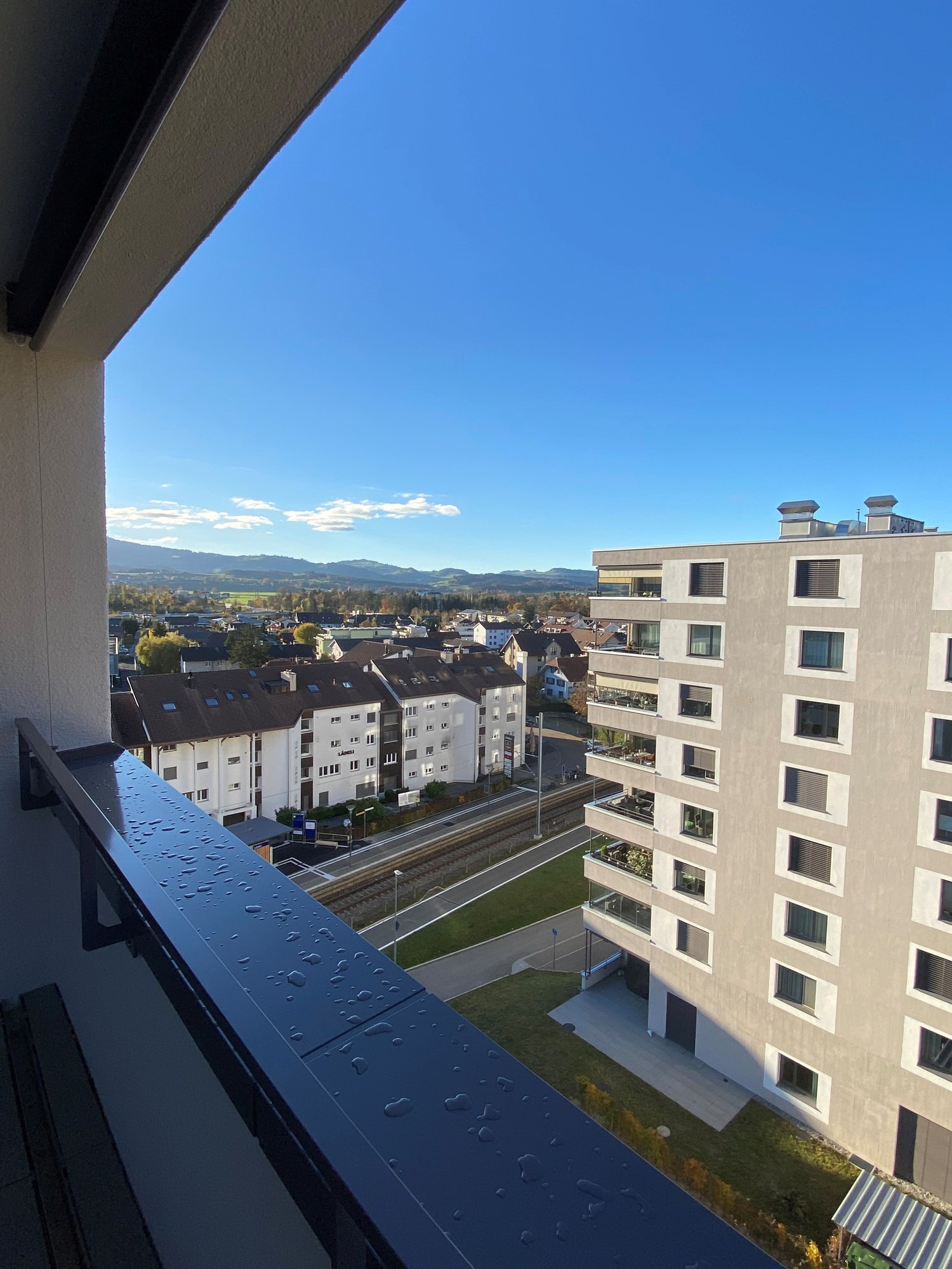 Balcony with railings, open to a view of city buildings, road, rail, trees, mountains