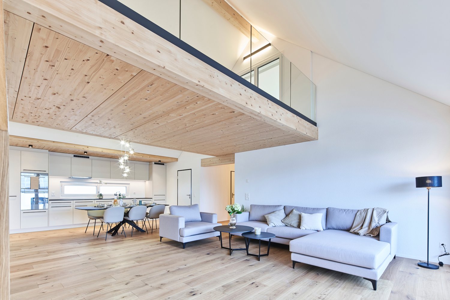 Spacious open-plan living area with wooden ceiling beams, large windows, modern kitchen with white cabinets, dining table with chairs, and a comfortable gray sofa set with coffee tables.
