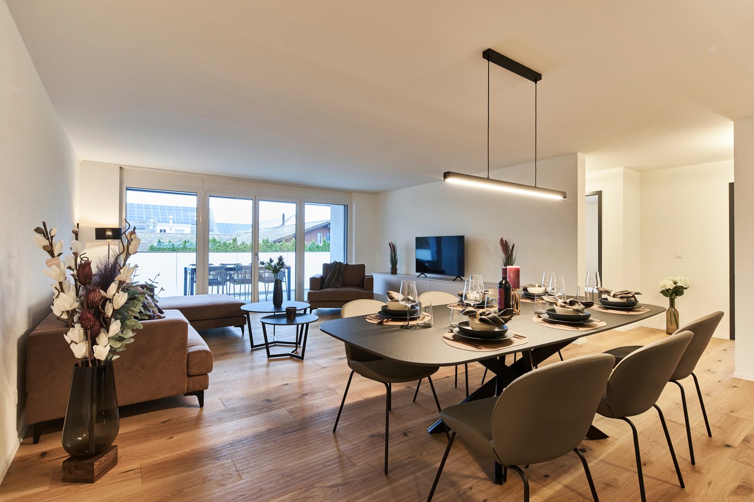 Open-concept living and dining area with large windows, hardwood floors, modern furniture including a sofa, coffee table, dining table and chairs, and a TV mounted on the wall. The room has a balcony or terrace visible through the windows.