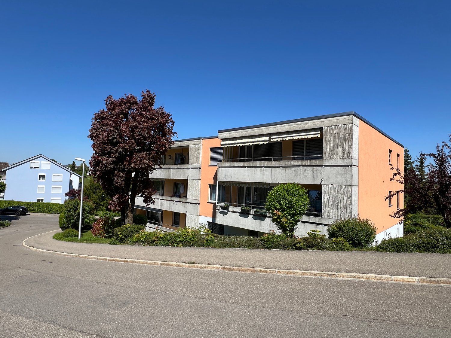 Modern multi-story building, orange exterior, balcony on second floor, small garden in front, car parked