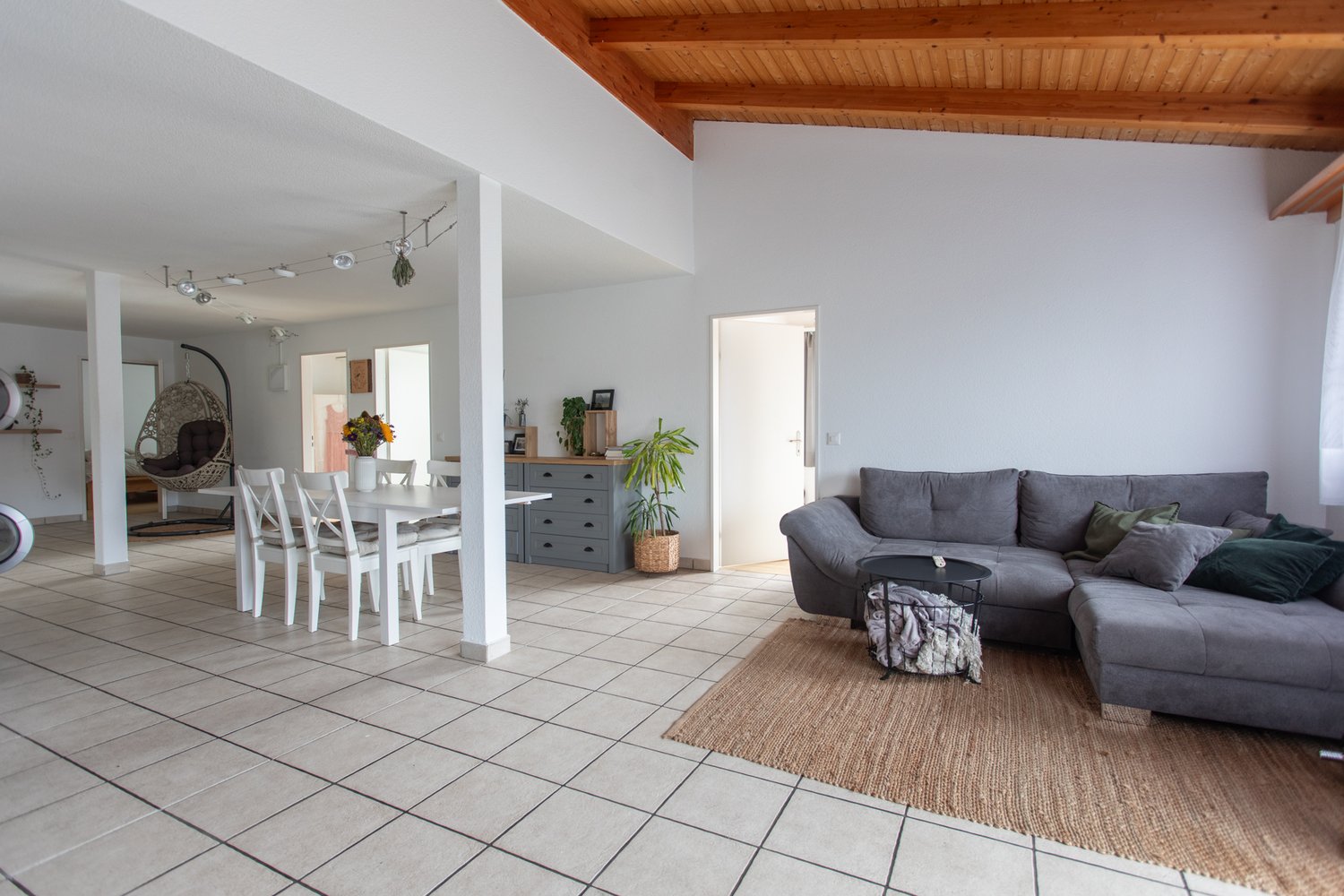 Open-concept living room and dining area, with a large gray sofa, wooden dining table and chairs, and a hanging chair. The room has tiled flooring, white walls, and a wooden ceiling with exposed beams. There is a balcony or terrace visible through the doo
