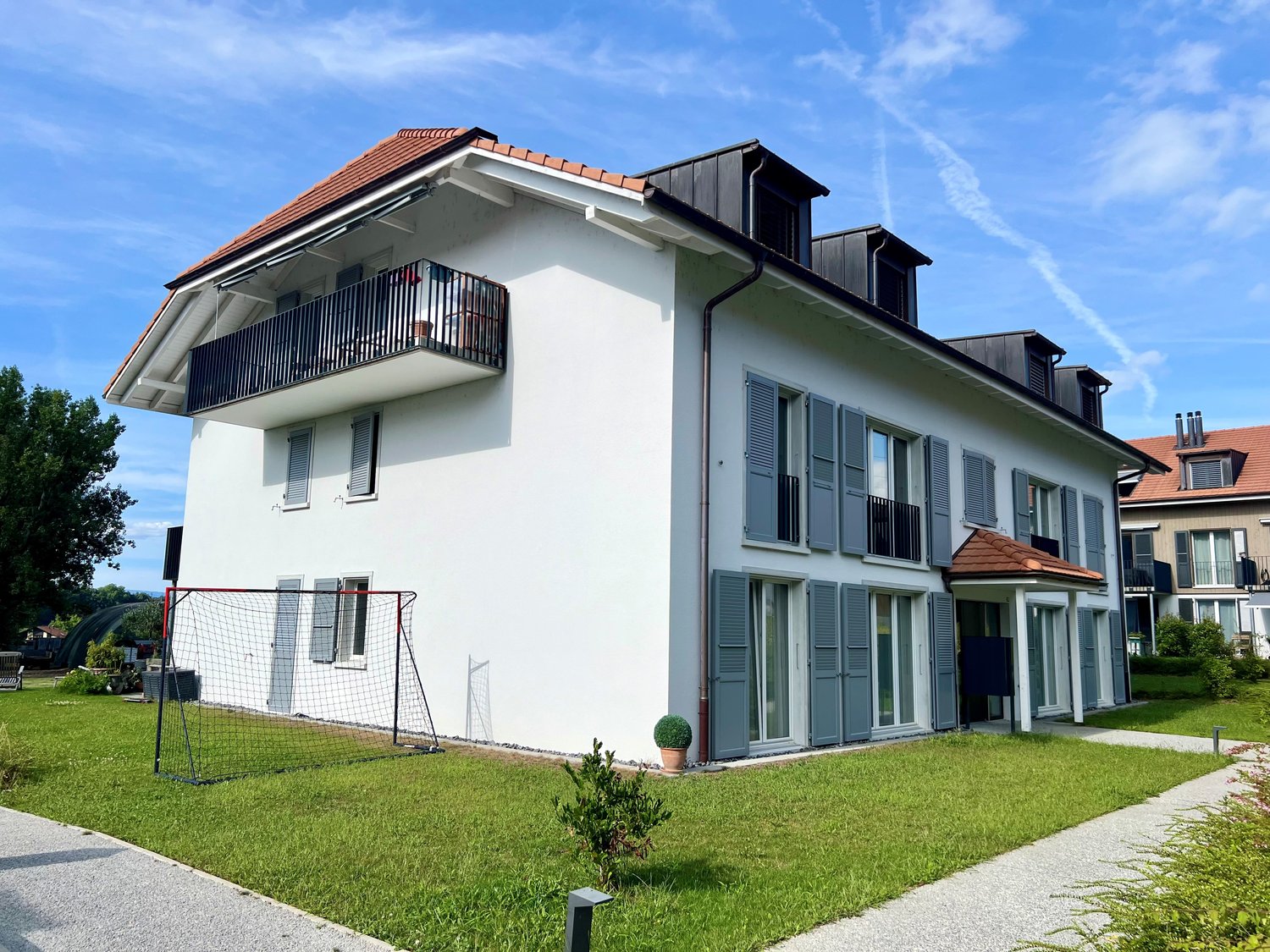 2-story apartment building with white exterior, red tile roof, balconies, and a grassy lawn with a soccer goal in the foreground.