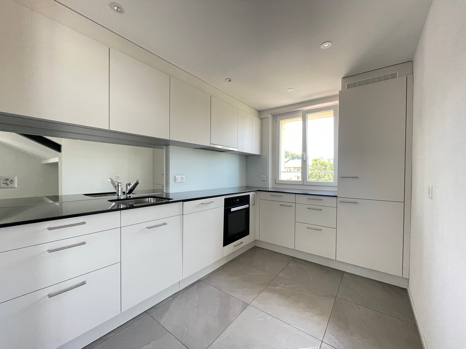 modern white kitchen, black countertops, grey floor, large window with outdoor view, built-in oven and dishwasher