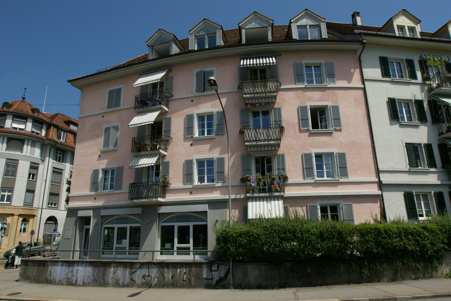 A building with many windows, balconies, and an awning. The building is painted pink and white and has multiple floors. A street light is in front of the building and bushes are next to it.